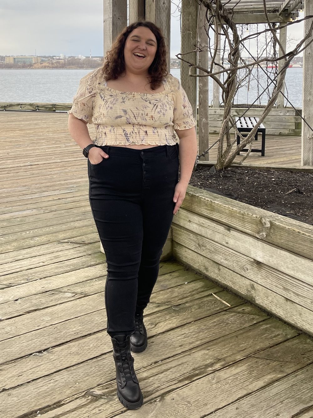 A person with curly hair, wearing a light-colored cropped blouse and black pants, stands on a wooden dock with a body of water in the background, smiling.