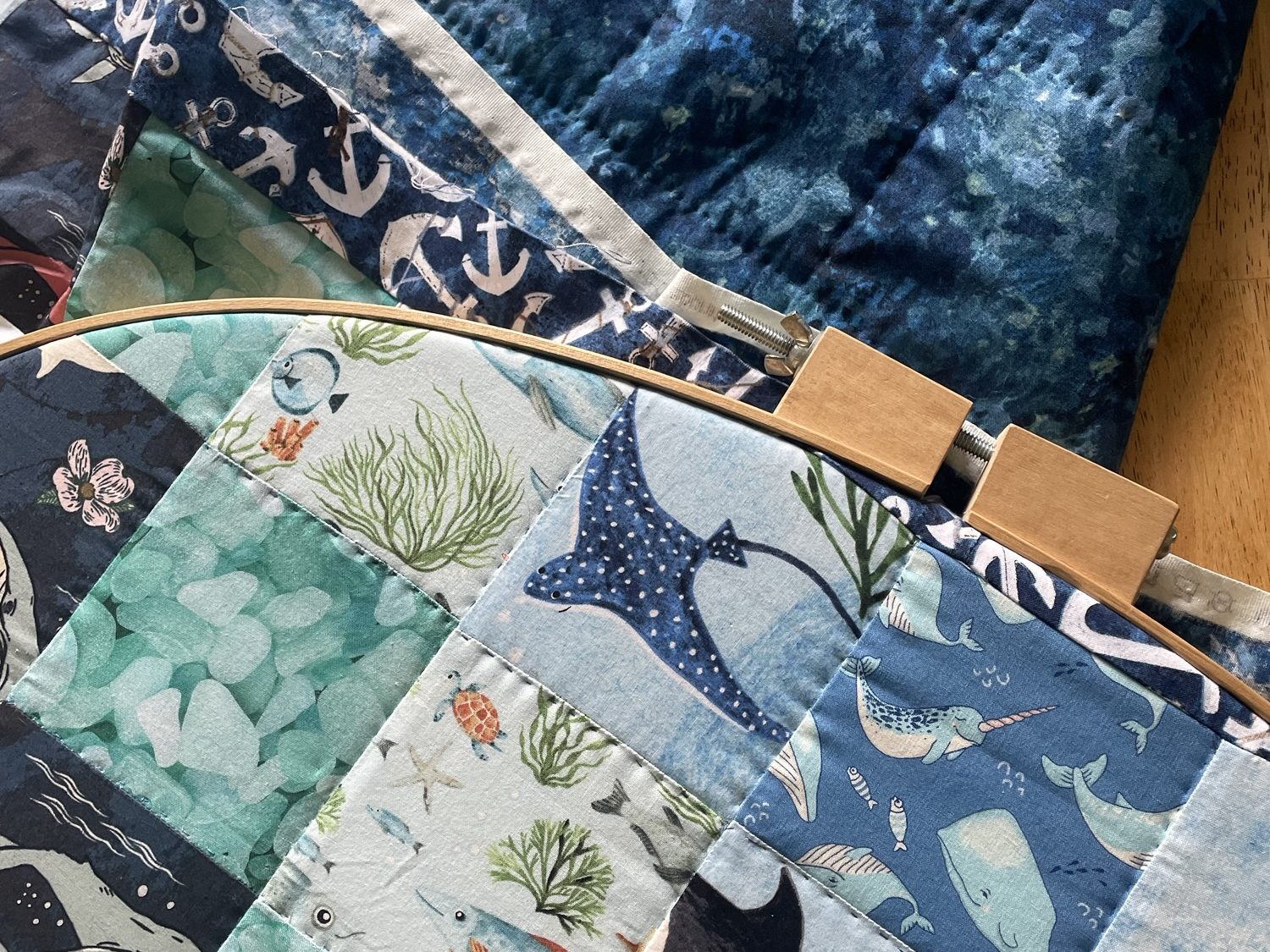 A close-up view of a colorful handstitched quilt featuring marine life illustrations such as fish, sea turtles, and seaweed, with a wooden embroidery hoop partially visible.